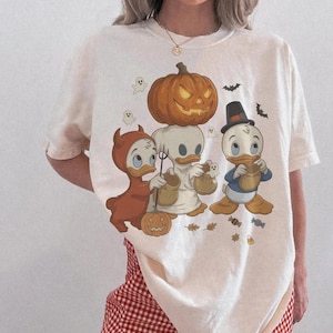 以下が含まれることがあります： ハロウィンの衣装を着た3羽のアヒルのキャラクターが描かれたクリーム色の半袖Tシャツ。1羽は赤い悪魔の衣装、1羽は幽霊の仮装、もう1羽はシルクハットをかぶっています。彼らの上には大きなジャック・オー・ランタンが配置されています。