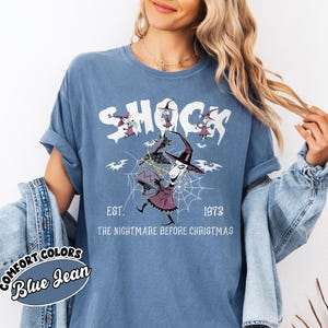 May include: Blue Comfort Colors t-shirt with a graphic from "The Nightmare Before Christmas." The design features the word "SHOCK" above a character in a witch hat and a spider web. The text "EST. 1973" and "THE NIGHTMARE BEFORE CHRISTMAS" are also included.
