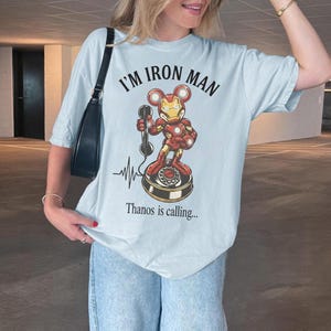 Op de afbeelding: Lichtblauw T-shirt met de tekst "I'M IRON MAN" en een stripfiguur Iron Man op een draaitelefoon. De zin "Thanos is calling..." staat eronder gedrukt. Het shirt heeft een casual stijl.