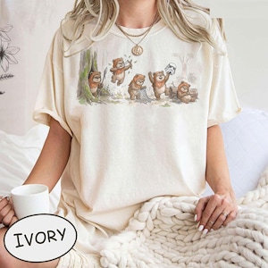 May include: Ivory-colored t-shirt featuring a whimsical illustration of Ewoks in a forest setting. The design includes several Ewoks with various props and a Stormtrooper helmet. The shirt has short sleeves and a relaxed fit.