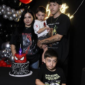 May include: A family poses for a photo at a birthday party. A two-tiered cake with a Spider-Man theme is on a table. Red and black balloons decorate the background. The family members are wearing Spider-Man themed shirts.