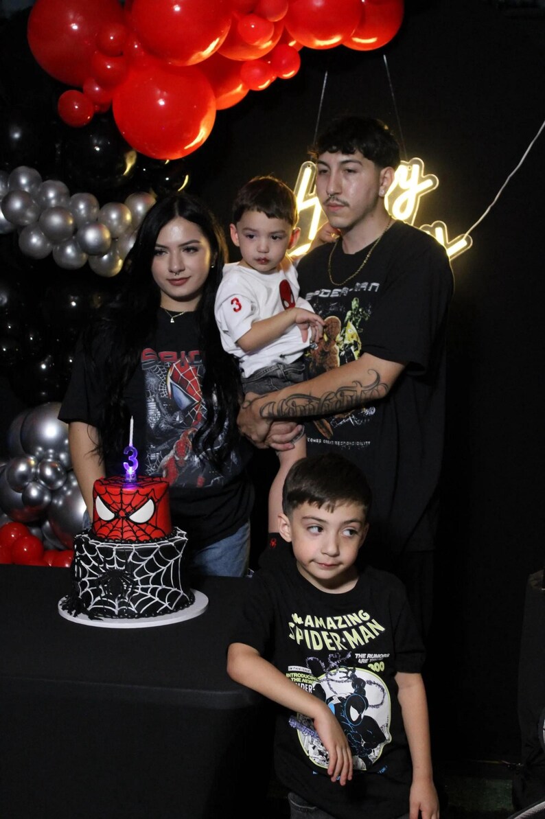 May include: A family poses for a photo at a birthday party. A two-tiered cake with a Spider-Man theme is on a table. Red and black balloons decorate the background. The family members are wearing Spider-Man themed shirts.