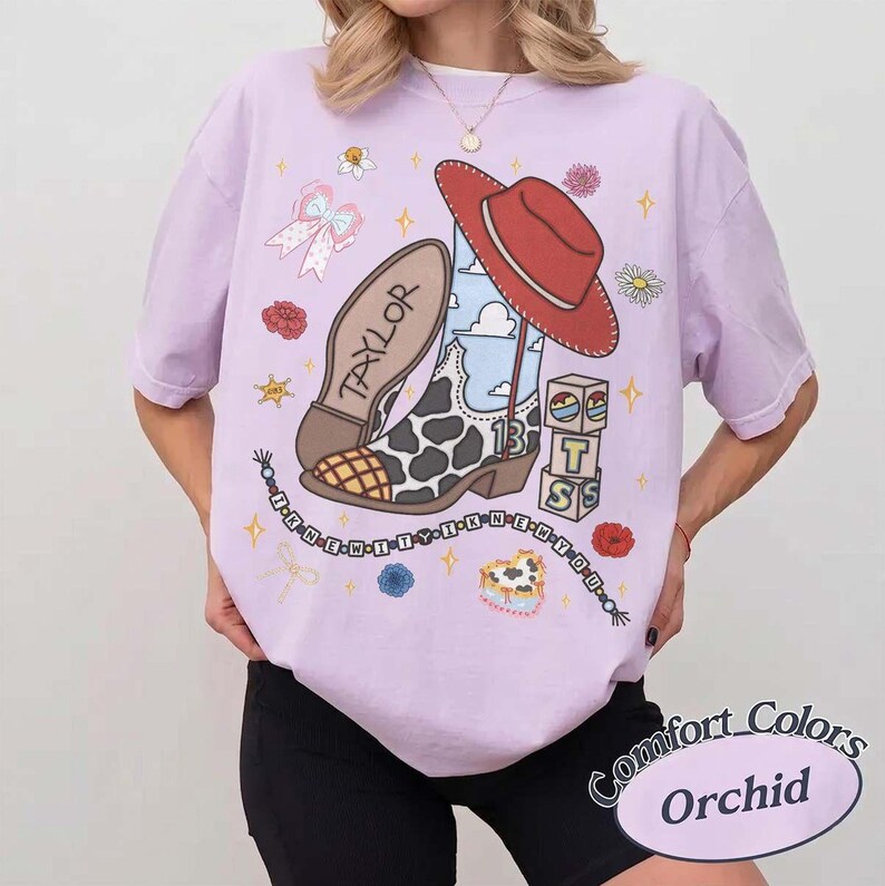 May include: A light purple t-shirt featuring a graphic print of a cowboy boot with a cow print pattern, a red hat, a bow, and blocks with the letters T, S. The text TAYLOR is on the boot sole, and a beaded string with text surrounds the design.