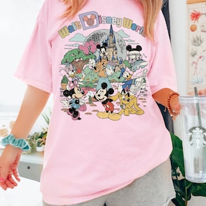 May include: Pink t-shirt featuring a colorful graphic of Disney characters and the text "Walt Disney World." The design includes Mickey Mouse, Minnie Mouse, Donald Duck, and other characters in a cartoon style. The shirt is a casual, oversized fit.