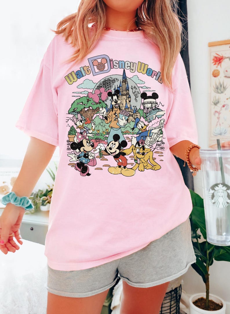 May include: Pink t-shirt featuring a colorful graphic of Disney characters and the text "Walt Disney World." The design includes Mickey Mouse, Minnie Mouse, Donald Duck, and other characters in a cartoon style. The shirt is a casual, oversized fit.