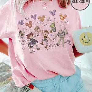 May include: Pink t-shirt featuring a graphic of Toy Story characters holding Mickey Mouse-shaped balloons. Characters include Mr. Potato Head, Jessie, Woody, Buzz Lightyear, and Alien. The shirt has the word "Blossom" on it.