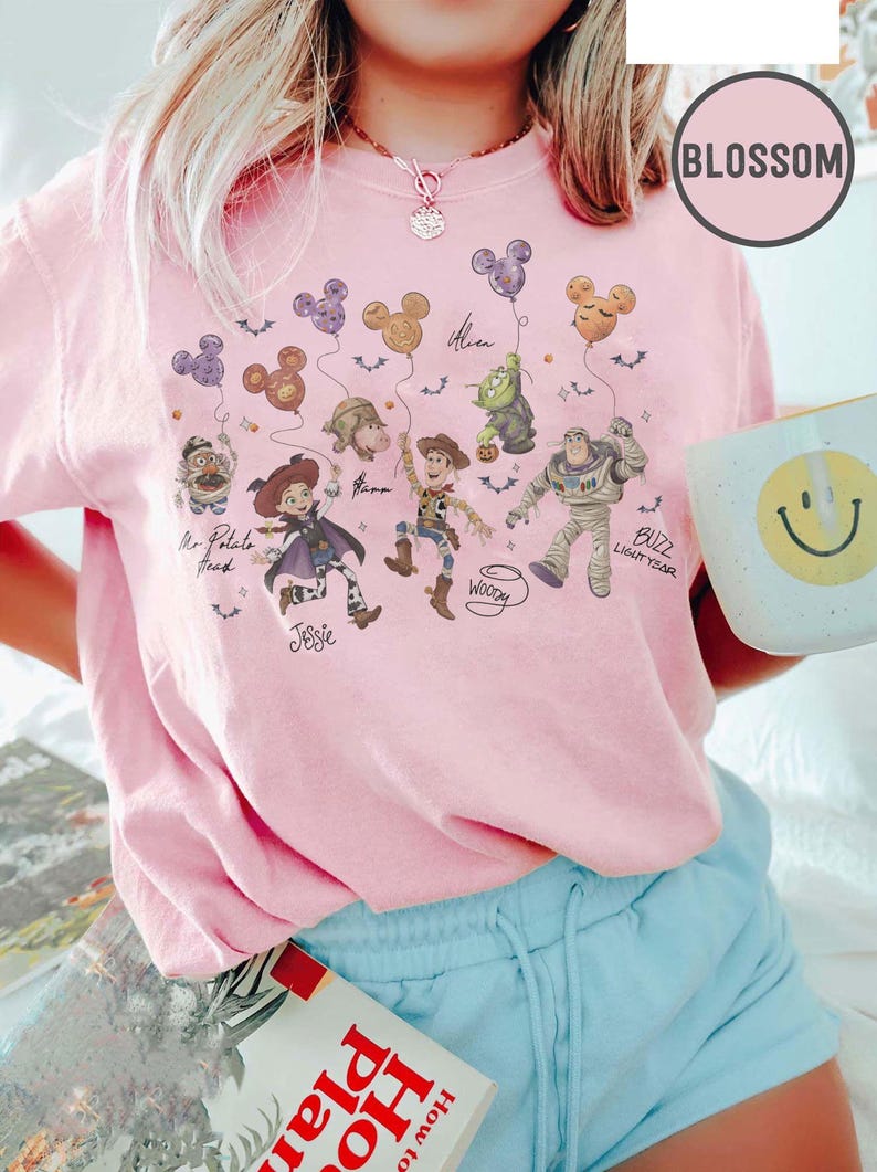 May include: Pink t-shirt featuring a graphic of Toy Story characters holding Mickey Mouse-shaped balloons. Characters include Mr. Potato Head, Jessie, Woody, Buzz Lightyear, and Alien. The shirt has the word "Blossom" on it.