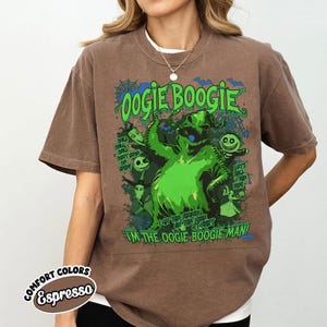 Op de afbeelding: Espresso-kleurig T-shirt met een groene Oogie Boogie-afbeelding met de tekst "Oogie Boogie" en "I'm the Oogie Boogie Man!". Het ontwerp bevat andere personages en zinnen uit de film. Het shirt is een comfort colors-stijl.