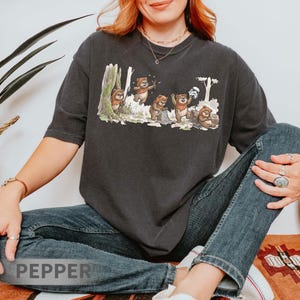 May include: Dark gray t-shirt featuring a whimsical illustration of Ewoks in a forest setting. The design includes several Ewoks interacting with each other, with a Stormtrooper helmet in the background. The word "PEPPER" is visible at the bottom.