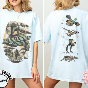 Pode incluir: Uma t-shirt azul claro com uma impress&atilde;o gr&aacute;fica de The Mandalorian e Grogu na frente, incluindo o texto "The Mandalorian and Grogu" e "This is the way". As costas da t-shirt exibem um alinhamento vertical de v&aacute;rios ve&iacute;culos Star Wars.