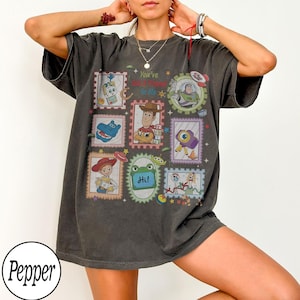 May include: A charcoal grey short-sleeve t-shirt featuring a graphic print of various Toy Story characters arranged in postage stamp-style frames. The text "You've Got A Friend In Me" is printed in the centre, surrounded by illustrations of Woody, Buzz Lightyear, Jessie, and other characters.