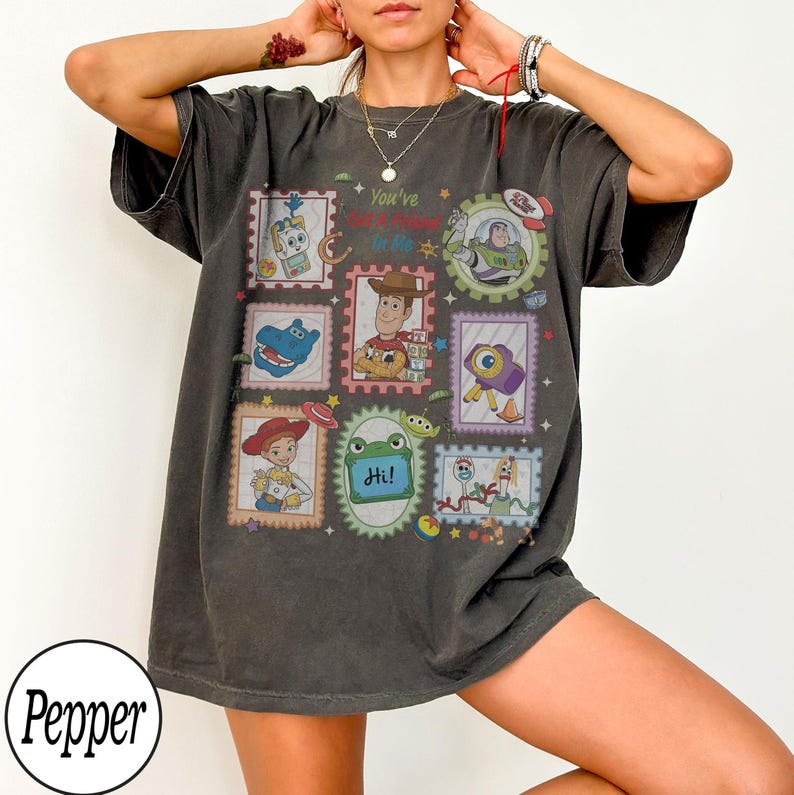 May include: A charcoal grey short-sleeve t-shirt featuring a graphic print of various Toy Story characters arranged in postage stamp-style frames. The text "You've Got A Friend In Me" is printed in the centre, surrounded by illustrations of Woody, Buzz Lightyear, Jessie, and other characters.