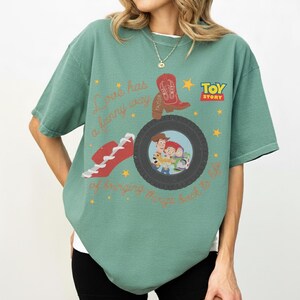 K&ouml;nnte beinhalten: Ein salbeigr&uuml;nes Kurzarm-T-Shirt mit einem Toy-Story-Motiv. Das Design zeigt einen roten Cowboystiefel, einen roten Sneaker, einen schwarzen Reifen, der Woody, Jessie und Buzz Lightyear einrahmt, sowie den Text Love has a funny way of bringing things back to life.