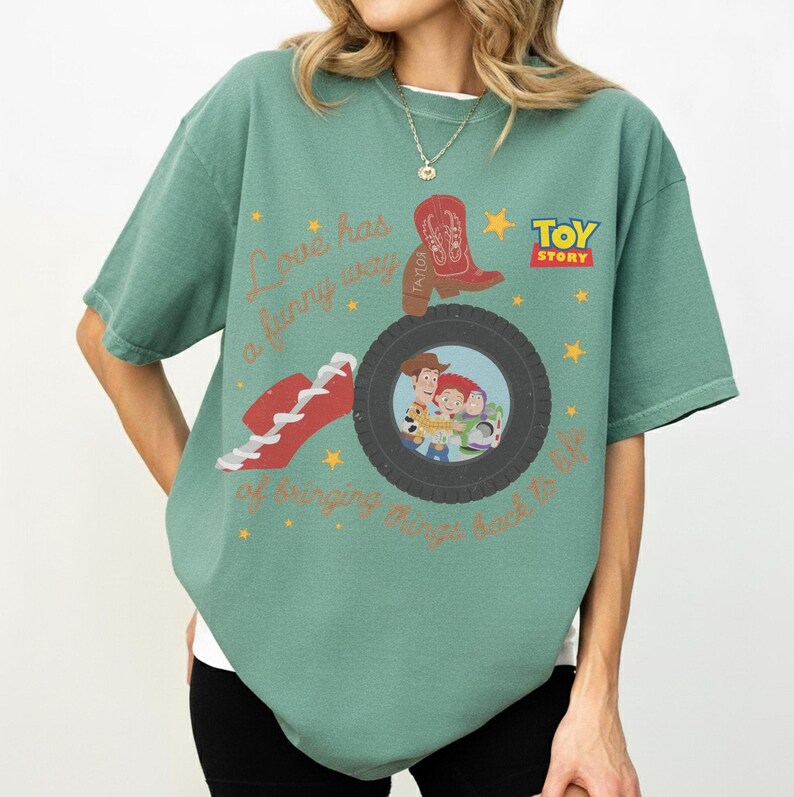 K&ouml;nnte beinhalten: Ein salbeigr&uuml;nes Kurzarm-T-Shirt mit einem Toy-Story-Motiv. Das Design zeigt einen roten Cowboystiefel, einen roten Sneaker, einen schwarzen Reifen, der Woody, Jessie und Buzz Lightyear einrahmt, sowie den Text Love has a funny way of bringing things back to life.