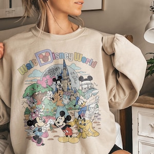 May include: Beige sweatshirt featuring a colorful graphic of Disney characters and the text "Walt Disney World." The design includes Mickey and Minnie Mouse, Donald Duck, and a castle. The sweatshirt has long sleeves.