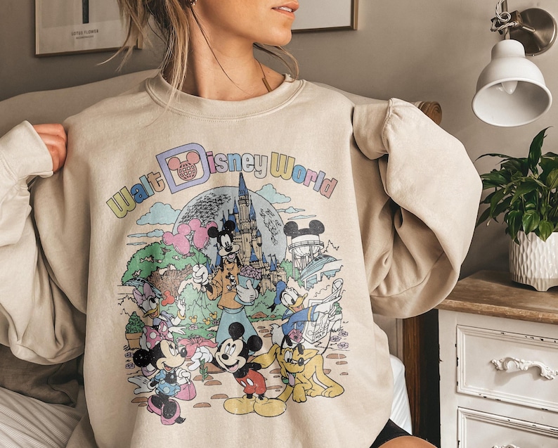 May include: Beige sweatshirt featuring a colorful graphic of Disney characters and the text "Walt Disney World." The design includes Mickey and Minnie Mouse, Donald Duck, and a castle. The sweatshirt has long sleeves.