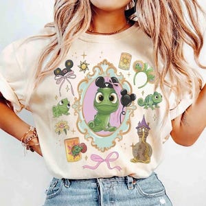Peut inclure: Un t-shirt couleur crème arborant un motif central représentant un caméléon vert dans un cadre bleu clair orné. Le design comprend de petites illustrations d'une haute tour, d'une lanterne jaune, d'une fleur et de serre-têtes à oreilles.