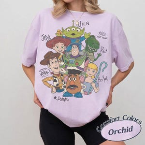 May include: An orchid-colored short-sleeve t-shirt featuring a graphic print of Toy Story characters including Woody, Buzz Lightyear, Jessie, Rex, Mr. Potato Head, Bo Peep, and an Alien, with their names handwritten in black ink around the illustration.