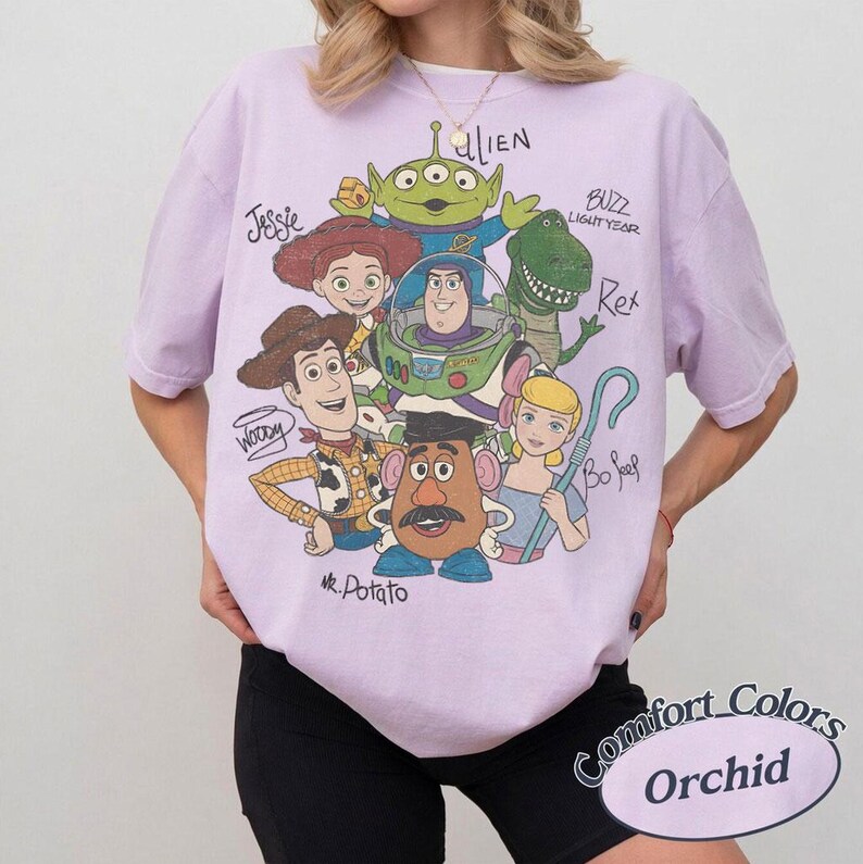 May include: An orchid-colored short-sleeve t-shirt featuring a graphic print of Toy Story characters including Woody, Buzz Lightyear, Jessie, Rex, Mr. Potato Head, Bo Peep, and an Alien, with their names handwritten in black ink around the illustration.