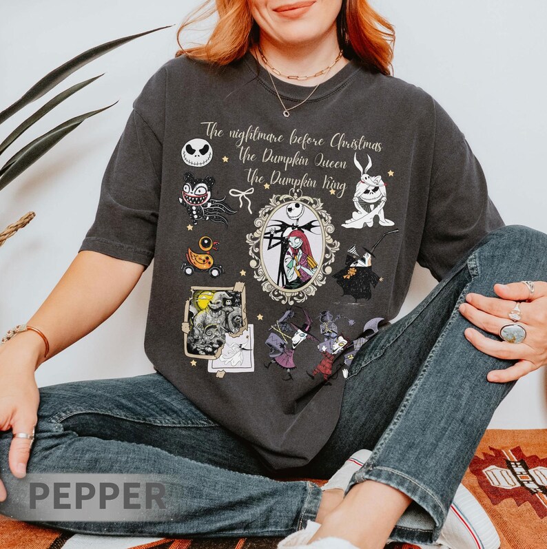 Pu&ograve; includere: T-shirt grigio scuro con personaggi e testo da "Nightmare Before Christmas". Il design include Jack Skellington, Sally e altri personaggi in stile vintage. Il testo recita "The Nightmare Before Christmas, The Pumpkin Queen, The Pumpkin King."