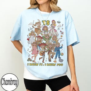 K&ouml;nnte beinhalten: Ein hellblaues T-Shirt mit einem Grafikdruck von mehreren Zeichentrickfiguren in verschiedenen Outfits, darunter ein Raumanzug und Cowgirl-Kleidung. Der Text "TS" steht oben und der Text "I KNEW IT, I KNEW YOU" ist unten aufgedruckt.