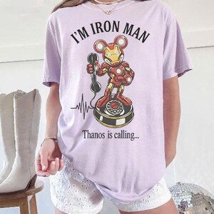 Op de afbeelding: Lichtpaars T-shirt met de tekst "I'M IRON MAN" boven een cartoon Iron Man-personage dat een telefoon vasthoudt. Onder het personage staat de tekst "Thanos is calling...". Er is ook een hartslaglijn aanwezig.