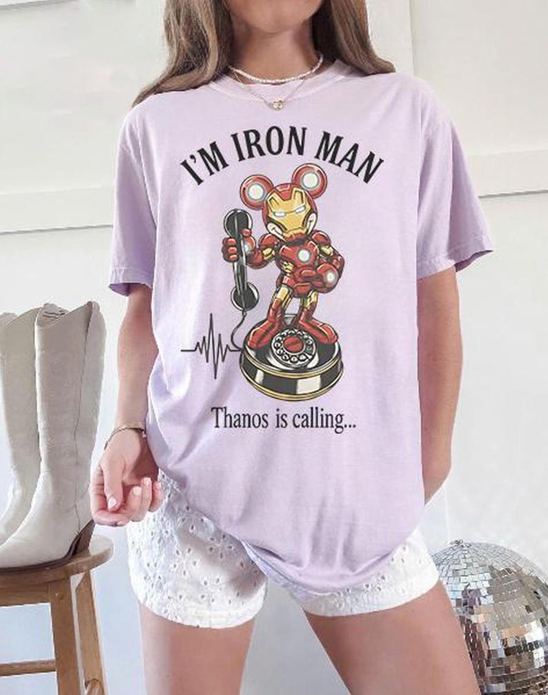 Op de afbeelding: Lichtpaars T-shirt met de tekst "I'M IRON MAN" boven een cartoon Iron Man-personage dat een telefoon vasthoudt. Onder het personage staat de tekst "Thanos is calling...". Er is ook een hartslaglijn aanwezig.
