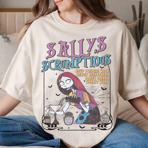 May include: Cream-colored t-shirt featuring a graphic of Sally from The Nightmare Before Christmas. The design includes the text "Sally's Scrumptious" and a recipe. The artwork is in shades of purple, blue, and black.