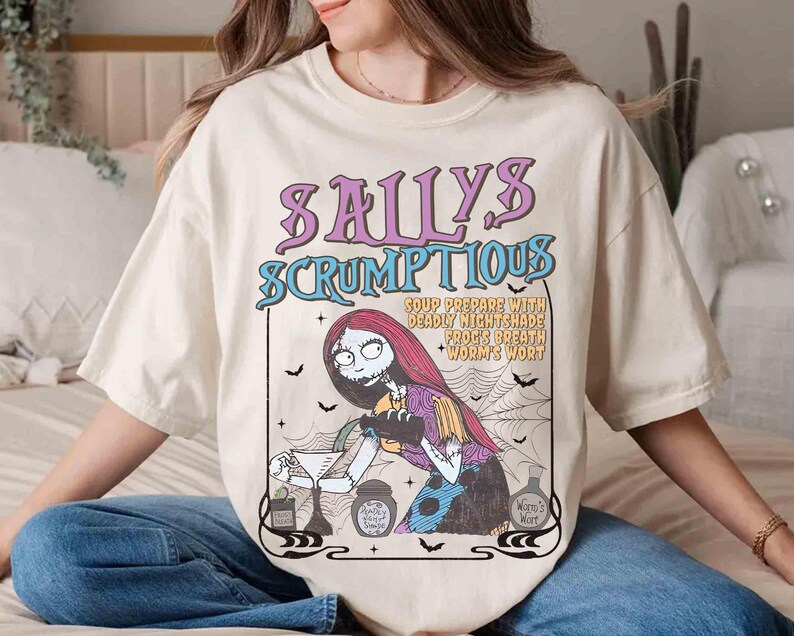May include: Cream-colored t-shirt featuring a graphic of Sally from The Nightmare Before Christmas. The design includes the text "Sally's Scrumptious" and a recipe. The artwork is in shades of purple, blue, and black.
