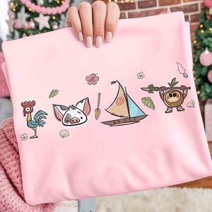 Pode incluir: Uma camisola rosa claro com uma impress&atilde;o gr&aacute;fica de um galo de desenho animado, uma cara de porco, um remo de madeira, um pequeno veleiro com vela estampada e uma personagem de coco com uma lan&ccedil;a, com pequenas flores e folhas tropicais.