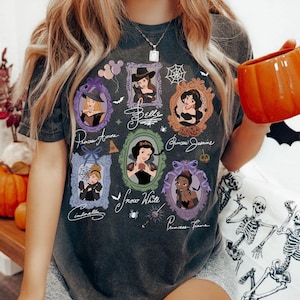 May include: Dark gray t-shirt featuring framed portraits of Disney princesses in Halloween costumes. The shirt includes the names Princess Aurora, Belle, Princess Jasmine, Cinderella, Snow White, and Princess Tiana. The design incorporates bats, spiders, and a witch's hat.