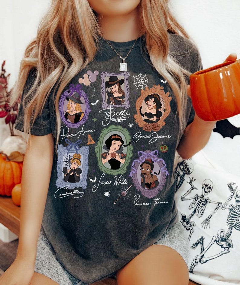 May include: Dark gray t-shirt featuring framed portraits of Disney princesses in Halloween costumes. The shirt includes the names Princess Aurora, Belle, Princess Jasmine, Cinderella, Snow White, and Princess Tiana. The design incorporates bats, spiders, and a witch's hat.