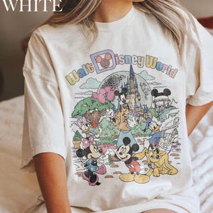 May include: Cream-colored t-shirt featuring a colorful graphic of Disney characters and the words "Walt Disney World." The graphic includes Mickey Mouse, Minnie Mouse, Donald Duck, and Goofy, with a castle and balloons.