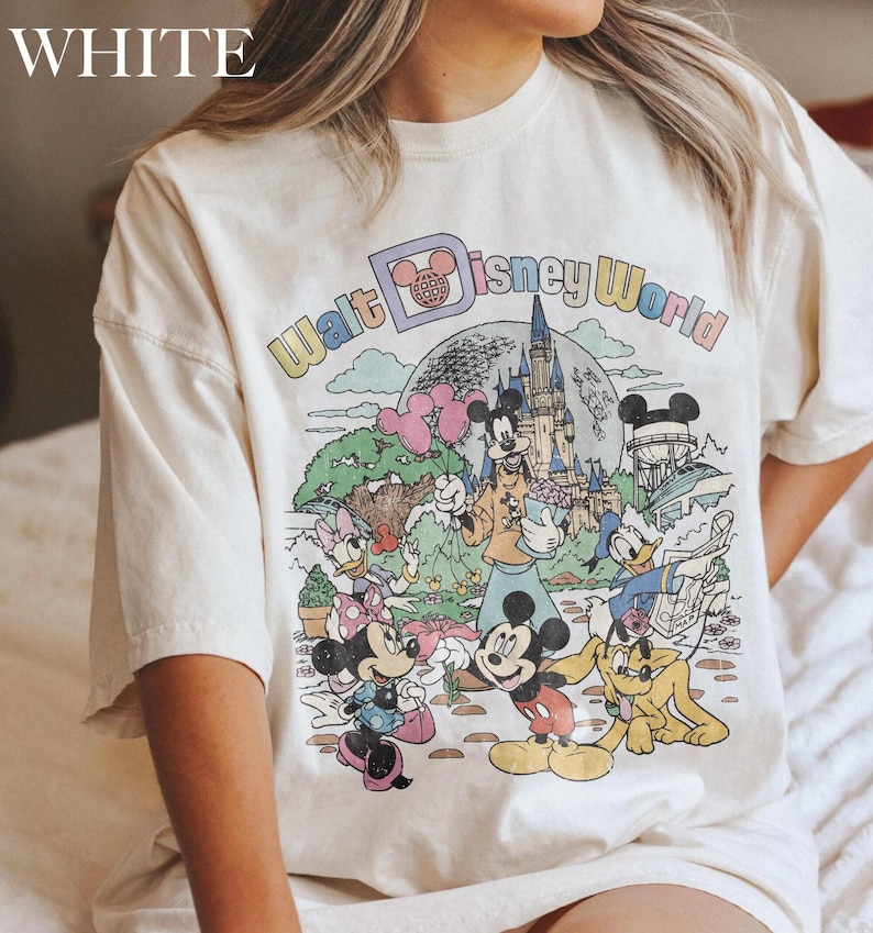 May include: Cream-colored t-shirt featuring a colorful graphic of Disney characters and the words "Walt Disney World." The graphic includes Mickey Mouse, Minnie Mouse, Donald Duck, and Goofy, with a castle and balloons.
