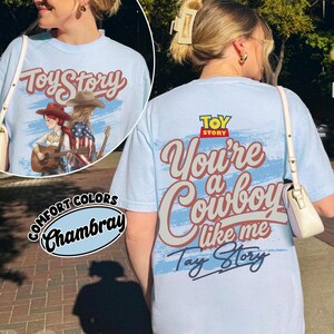 May include: A light blue t-shirt featuring a graphic print on the back with the text Toy Story, You're a Cowboy like me, and Tay Story in a script font. The design includes a stylised illustration of two figures with guitars and cowboy hats.