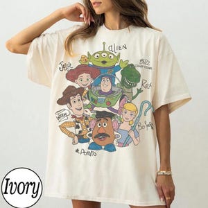 May include: An ivory short-sleeve t-shirt featuring a graphic print of Toy Story characters including Woody, Buzz Lightyear, Jessie, Rex, Mr. Potato Head, Bo Peep, and an Alien. The names of each character are printed in black script next to them.