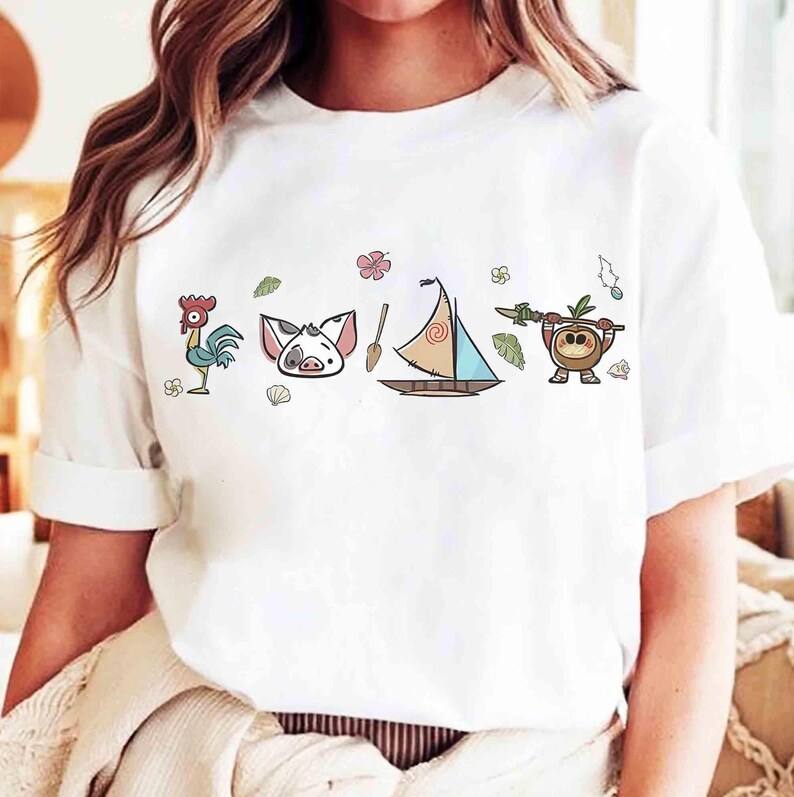 Pode incluir: Uma t-shirt branca de manga curta com uma impress&atilde;o gr&aacute;fica horizontal de um galo de desenho animado, uma cara de porco, um remo de madeira, um veleiro com vela estampada e uma pequena criatura de coco com uma lan&ccedil;a, decorada com flores.