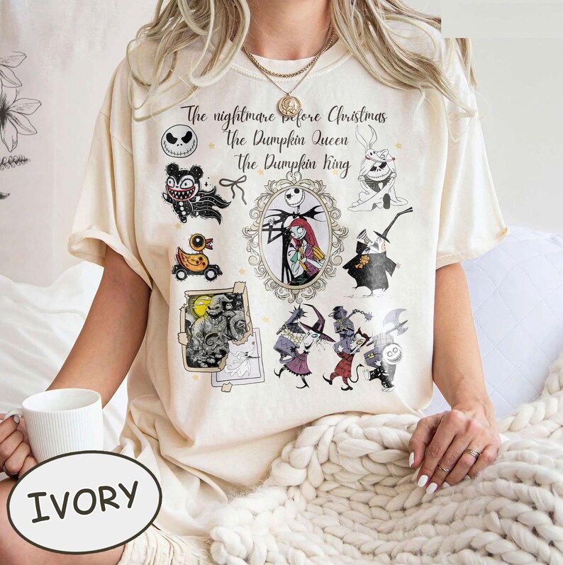 Pu&ograve; includere: T-shirt color avorio con una grafica di "Nightmare Before Christmas". Il design include personaggi come Jack Skellington, Sally e altri, con il testo "The Nightmare Before Christmas, The Pumpkin Queen, The Pumpkin King."