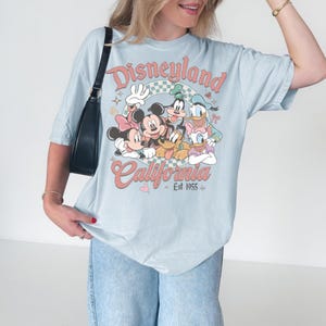 May include: Light blue t-shirt featuring a Disneyland California graphic with Mickey Mouse, Minnie Mouse, Goofy, Donald Duck, and Daisy Duck. The graphic is in a retro style with the text "Disneyland California Est 1955".