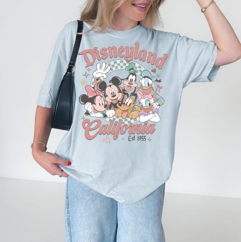 May include: Light blue t-shirt featuring a Disneyland California graphic with Mickey Mouse, Minnie Mouse, Goofy, Donald Duck, and Daisy Duck. The graphic is in a retro style with the text "Disneyland California Est 1955".