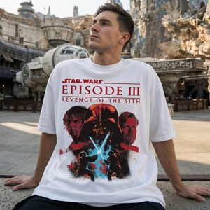 May include: A white short-sleeved t-shirt featuring a graphic print of Star Wars Episode III Revenge of the Sith. The design includes the movie title in red text above illustrations of Anakin Skywalker, Darth Vader, and Obi-Wan Kenobi.