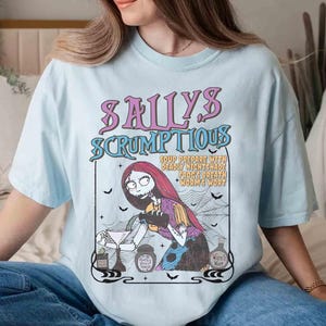 May include: Light blue t-shirt featuring a graphic of Sally from The Nightmare Before Christmas. The graphic reads "Sally's Scrumptious" with a recipe for soup. The design includes bats, a spider web, and a potion bottle.