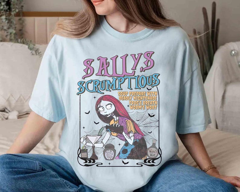 May include: Light blue t-shirt featuring a graphic of Sally from The Nightmare Before Christmas. The graphic reads "Sally's Scrumptious" with a recipe for soup. The design includes bats, a spider web, and a potion bottle.