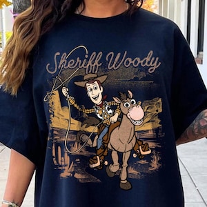 Pode incluir: Camiseta azul marinho com um desenho do Xerife Woody de Toy Story montado a cavalo. O texto "Sheriff Woody" est&aacute; acima do desenho em uma fonte brilhante. O design &eacute; ambientado em uma paisagem des&eacute;rtica.