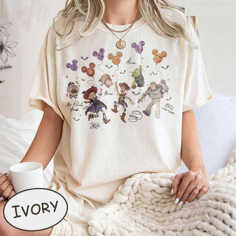 May include: Ivory-colored t-shirt featuring a colorful graphic of cartoon characters holding Mickey Mouse-shaped balloons. The characters include Mr. Potato Head, Jessie, Woody, Buzz Lightyear, and others. The word "IVORY" is printed on a white oval.
