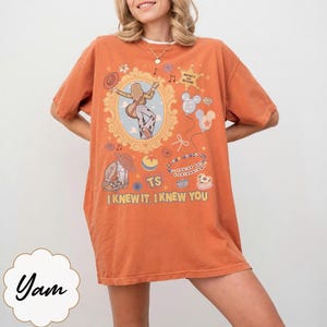 May include: An orange short-sleeve t-shirt featuring a graphic print with a central oval frame containing a figure playing a guitar, surrounded by various icons including balloons, a cowboy boot, and text reading "INFINITY AND BEYOND", "YOU'VE GOT A FRIEND IN ME", "TS", and "I KNEW IT, I KNEW YOU".