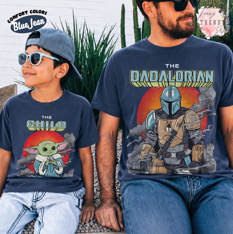 May include: Two navy blue t-shirts featuring graphic prints. The smaller shirt displays a green character with the text THE CHILD, while the larger shirt shows a masked armored figure with the text THE DADALORIAN. Both shirts feature a red sun background.