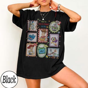 May include: A black short-sleeve t-shirt featuring a graphic print of various cartoon characters from Toy Story arranged in colourful postage stamp-style frames. The text "You've Got a Friend In Me" is printed in the centre above the character Woody.