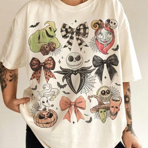 May include: An off-white short-sleeve t-shirt featuring a graphic print of various characters and bows. The design includes Oogie Boogie, Sally, Jack Skellington, Zero on a pumpkin, and Lock, Shock, and Barrel, interspersed with spiderwebs and bats.