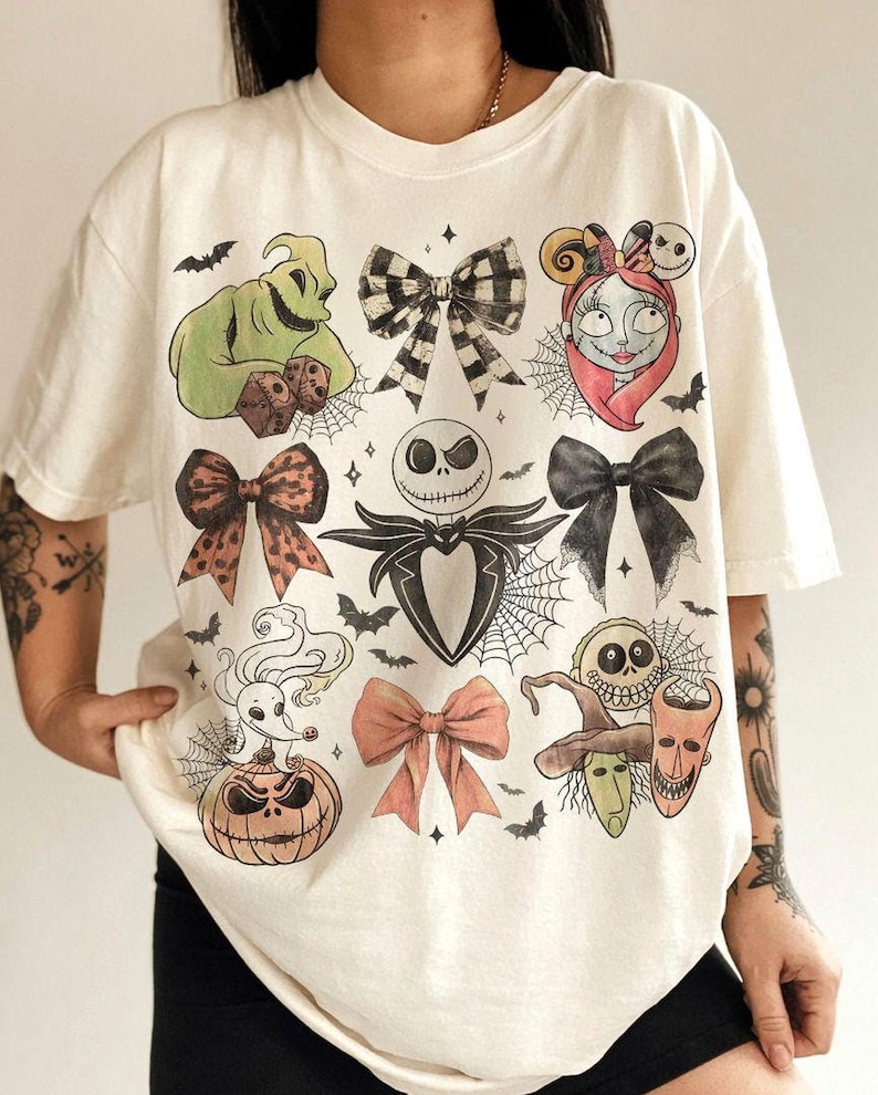 May include: An off-white short-sleeve t-shirt featuring a graphic print of various characters and bows. The design includes Oogie Boogie, Sally, Jack Skellington, Zero on a pumpkin, and Lock, Shock, and Barrel, interspersed with spiderwebs and bats.